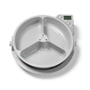 Ein grau automatischer Haustierfütterer mit geöffnetem Deckel, der drei interne Futterkammern zeigt, die durch einen zentralen Rotationsmechanismus getrennt sind. Ein digitales Bedienfeld mit LCD-Display ist in der oberen rechten Ecke sichtbar.